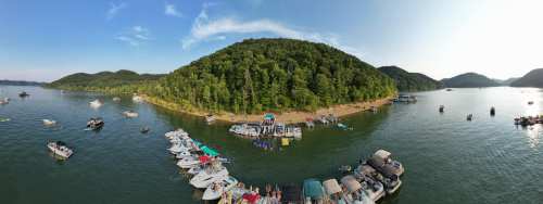 A panoramic view of a lake surrounded by green hills, with boats docked and people enjoying the water.