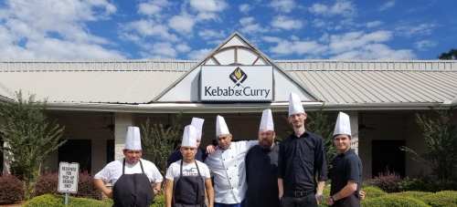 A group of six chefs in white hats and black uniforms stand outside Kebab & Curry restaurant under a blue sky.