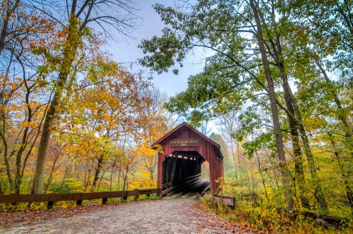 A red covered bridge surrounded by vibrant autumn foliage and trees, leading into a misty landscape.