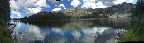 A serene lake surrounded by mountains and trees, reflecting clouds and blue sky in calm waters.