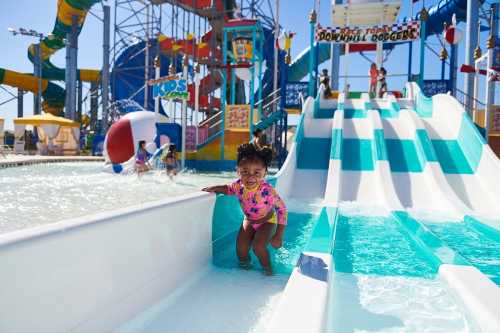 A young child in a colorful swimsuit climbs up a water slide at a fun water park, with splashes and other kids in the background.