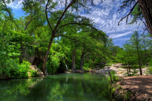 Lush green trees surround a tranquil river under a blue sky with fluffy clouds, creating a serene natural landscape.