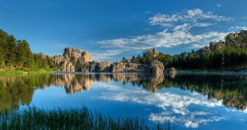 A serene lake surrounded by rocky formations and lush trees, reflecting a clear blue sky with scattered clouds.