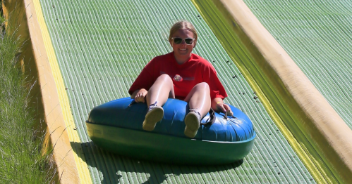 A person in a red shirt slides down a green and yellow tubing slope, smiling while sitting on a blue inflatable tube.