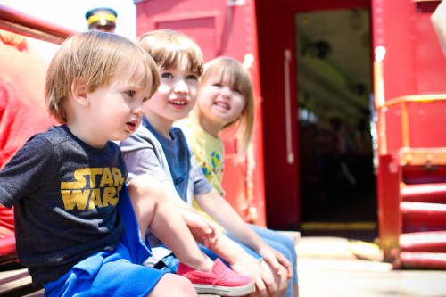 Three children sitting on a train bench, smiling and enjoying a sunny day.