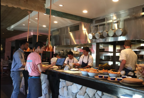 A busy kitchen with chefs preparing food and staff interacting at the counter, featuring pots and bowls of ingredients.