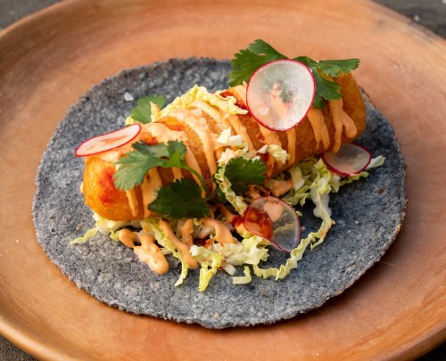 A blue corn tortilla topped with a crispy roll, lettuce, radish slices, and drizzled with sauce and cilantro.