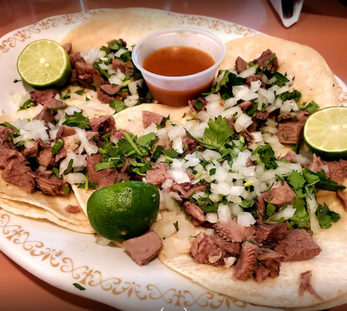 A plate of tacos filled with chopped meat, onions, and cilantro, served with lime wedges and a small cup of sauce.