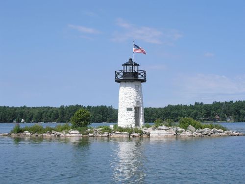 A white lighthouse with a black top and American flag stands on a small island surrounded by calm water and greenery.