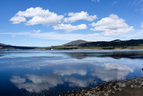A serene lake reflecting clouds and mountains under a blue sky, with rocky shoreline in the foreground.