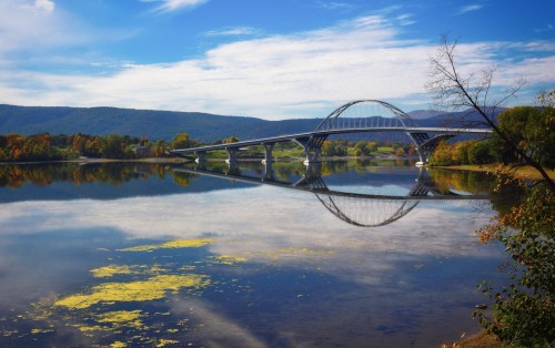 A modern bridge spans a calm river, reflecting the blue sky and surrounding trees in the water below.