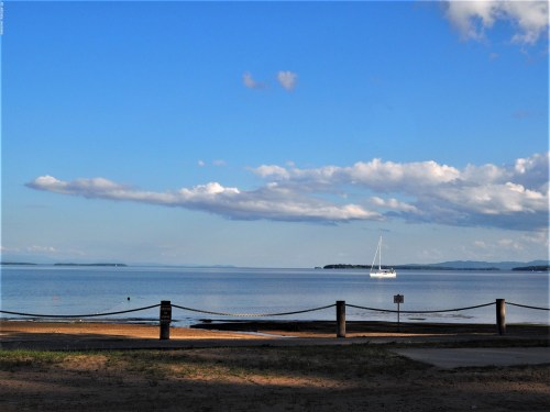 A serene beach scene with a sailboat on calm waters under a blue sky with fluffy clouds.