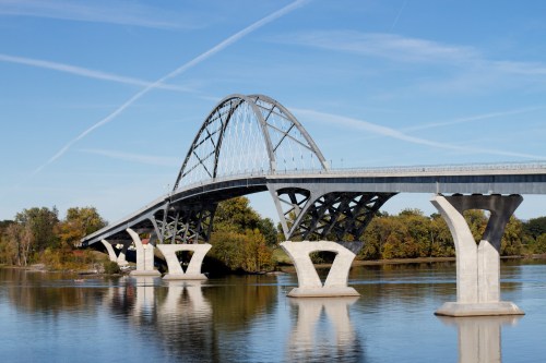 A modern arch bridge spans a river, supported by distinctive concrete pillars, with trees lining the shore under a clear blue sky.