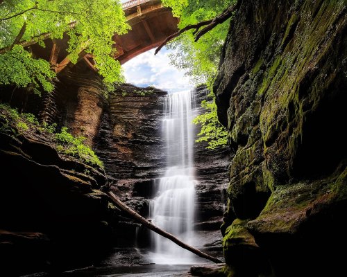 A serene waterfall cascades down rocky cliffs, framed by lush greenery and a bridge overhead.