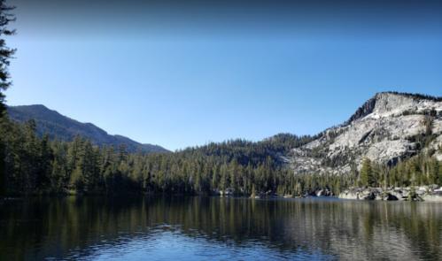 A serene lake surrounded by tall trees and mountains under a clear blue sky. Reflections shimmer on the water's surface.