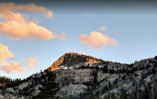 A rocky mountain peak bathed in golden sunlight, surrounded by trees and a blue sky with fluffy clouds.