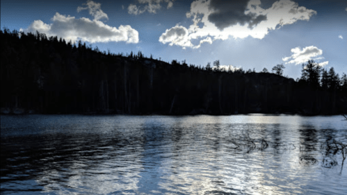 A serene lake reflects the sky, surrounded by dark trees and a dramatic cloud-filled horizon.