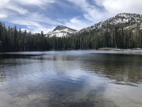 A serene lake surrounded by evergreen trees and snow-capped mountains under a partly cloudy sky.