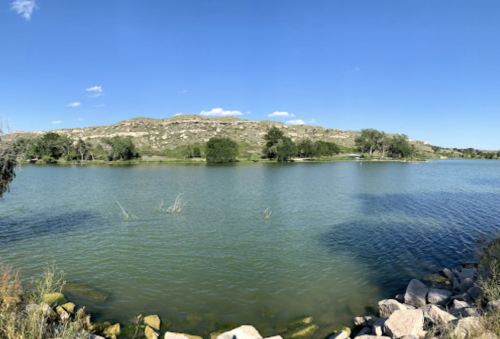 A serene lake surrounded by greenery and hills under a clear blue sky. Rocks line the shore.