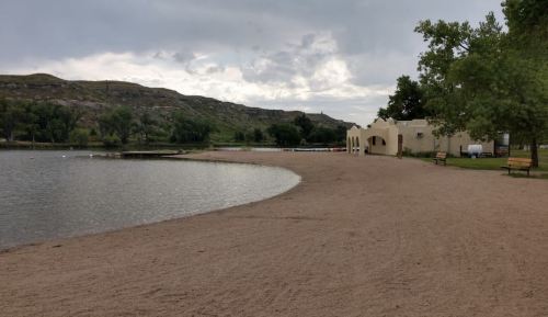 A sandy beach by a calm lake, with trees and a building in the background under a cloudy sky.