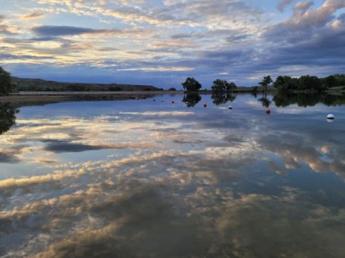A serene lake at sunset, reflecting colorful clouds and trees, with buoys floating on the calm water.