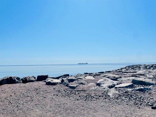 A calm sea under a clear blue sky, with a distant ship and rocky shoreline in the foreground.