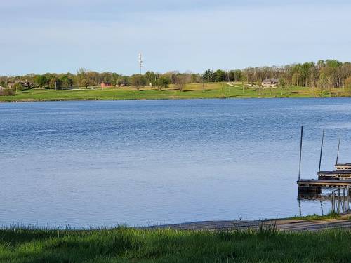 A calm lake scene with green hills, a few houses in the background, and a wooden dock in the foreground.