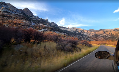 A scenic view of a winding road alongside rocky mountains and grassy fields under a clear blue sky.