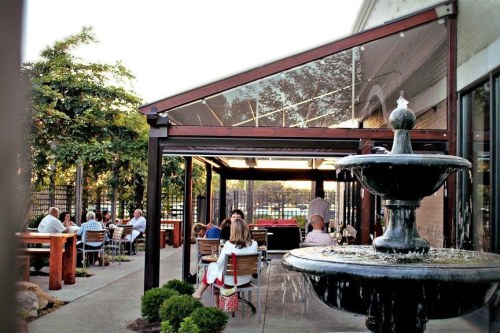 Outdoor dining area with a fountain, surrounded by greenery and people enjoying their meals under a glass-roofed structure.