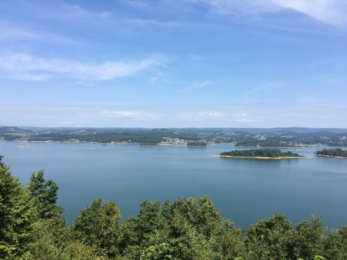 A serene lake surrounded by green trees and distant hills under a clear blue sky.
