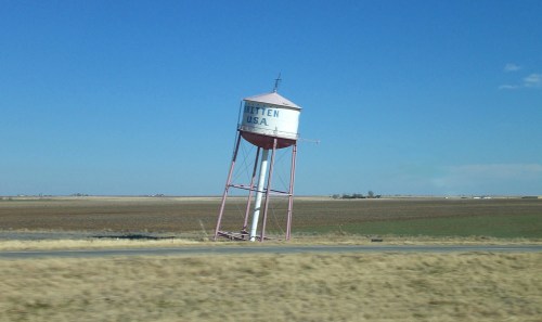 A leaning water tower with "Ritten, USA" painted on it, set against a clear blue sky and open fields.