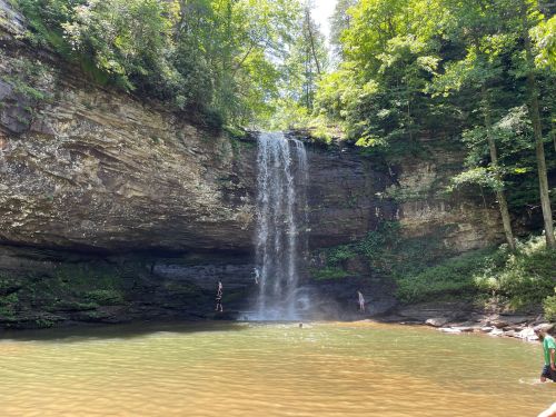 A serene waterfall cascades into a calm pool, surrounded by lush greenery and rocky cliffs. Two people are near the water.