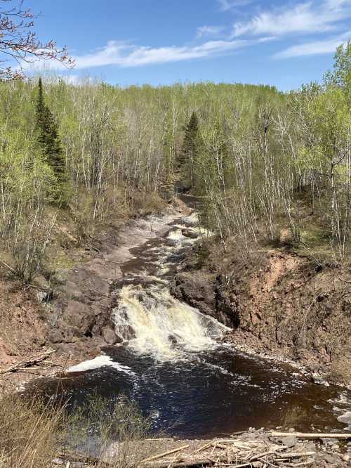 A serene waterfall cascades through rocky terrain, surrounded by lush green trees and a clear blue sky.