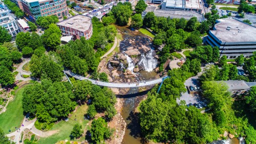 Aerial view of a park with a suspension bridge over a stream, surrounded by trees and buildings. Waterfall visible nearby.