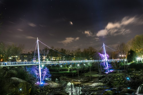 A illuminated suspension bridge at night, surrounded by trees and a river, under a starry sky.