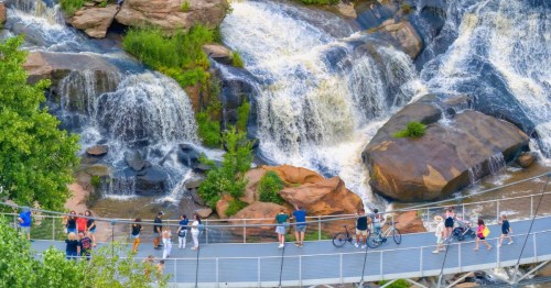 A scenic view of a waterfall with people walking on a bridge nearby, surrounded by lush greenery and rocky terrain.