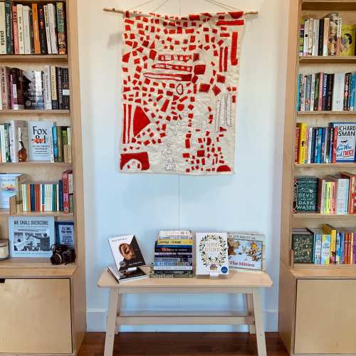 A cozy room with bookshelves filled with books, a textile wall hanging, and a small table displaying more books.