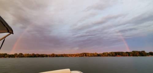 A serene lake scene with a double rainbow arching over tree-lined shores under a cloudy sky.
