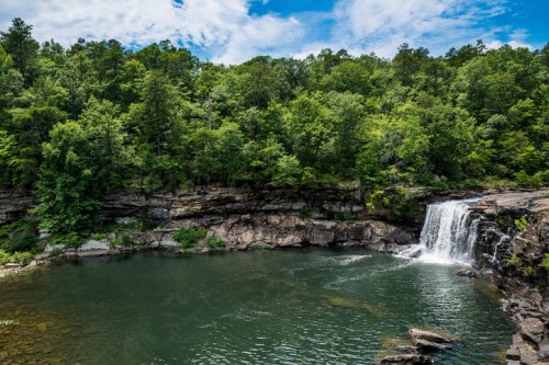 A serene waterfall cascades into a tranquil pool, surrounded by lush green trees and rocky cliffs under a blue sky.