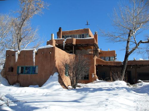 A southwestern-style adobe house covered in snow, surrounded by bare trees under a clear blue sky.