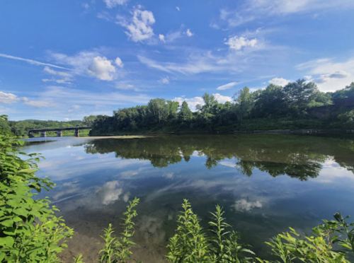A serene river scene with lush greenery and a blue sky reflected in the calm water, under fluffy white clouds.
