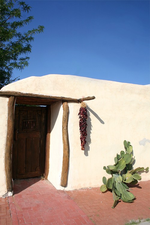 A rustic adobe building with a wooden door, hanging red chilies, and a cactus plant nearby under a clear blue sky.