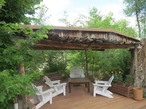 A rustic outdoor seating area with white chairs and a wooden table, surrounded by greenery and flowers.
