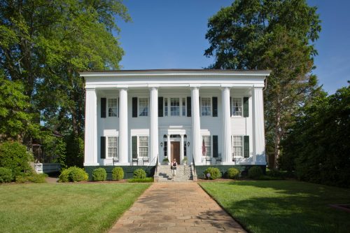 A large white historic house with columns, surrounded by greenery and a clear blue sky.