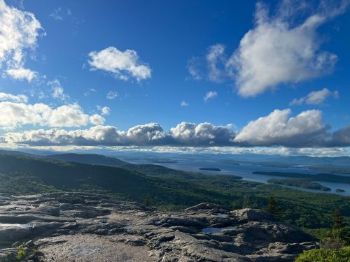 A panoramic view of lush green mountains and a serene lake under a bright blue sky with fluffy clouds.
