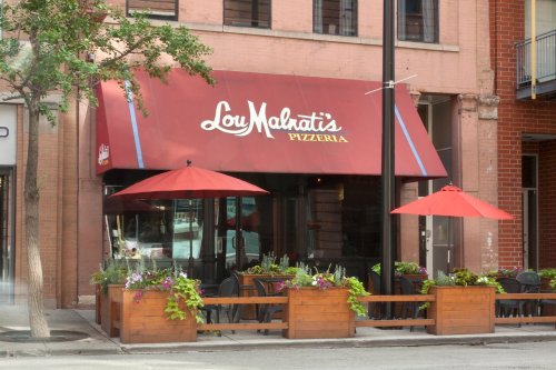 Exterior of Lou Malnati's Pizzeria with a red awning, outdoor seating, and planters filled with flowers.