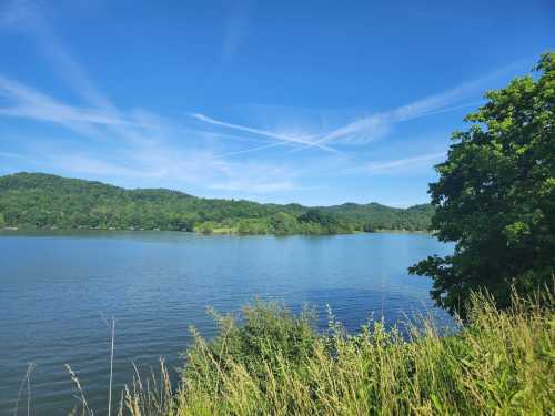 A serene lake surrounded by lush green hills under a clear blue sky with wispy clouds.