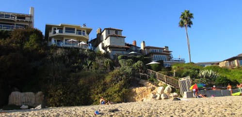 Beach scene with upscale homes on a hillside, palm trees, and people relaxing on the sand. Clear blue sky.