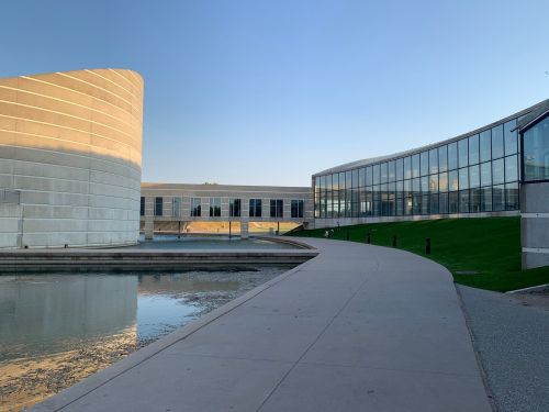 Modern architectural buildings beside a calm water feature, with a curved pathway and green grass in the foreground.