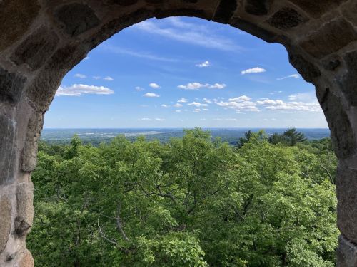 View through a stone archway, showcasing lush green trees and a blue sky with scattered clouds in the distance.